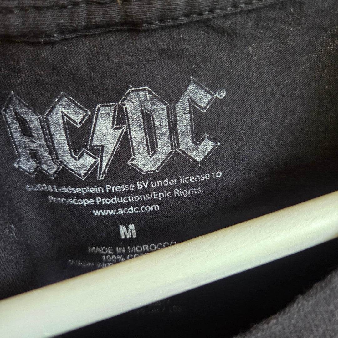 Close-up of a shirt tag featuring the AC/DC logo in white, along with licensing and manufacturing details, and a size M label below. The Upcycled Breastfeeding (Nursing) & Pumping Black Leopard AC/DC Band t-shirt M (100587) is designed for comfort with eco-friendly materials. A hanger is visible beneath the collar.