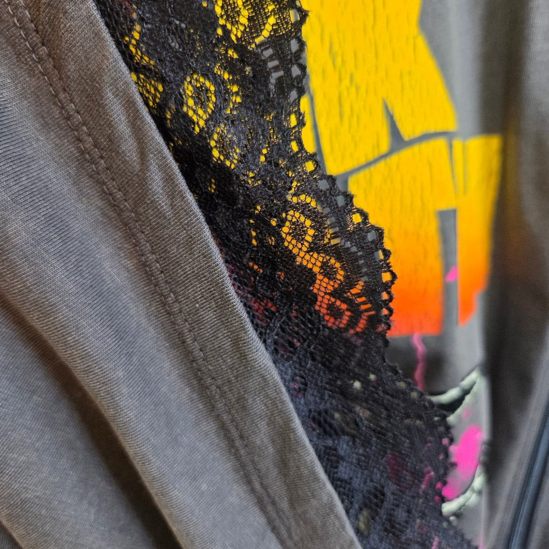 A close-up of the Primark Upcycled Breastfeeding (Nursing) & Pumping Grey/Orange Lace Black Sabbath Oversized Band T-shirt (Size S) reveals delicate black lace layered over grey fabric, with a hint of the vibrant orange and yellow band graphic beneath—highlighting the stylish and detailed design featured in this affordable maternity and nursing top.