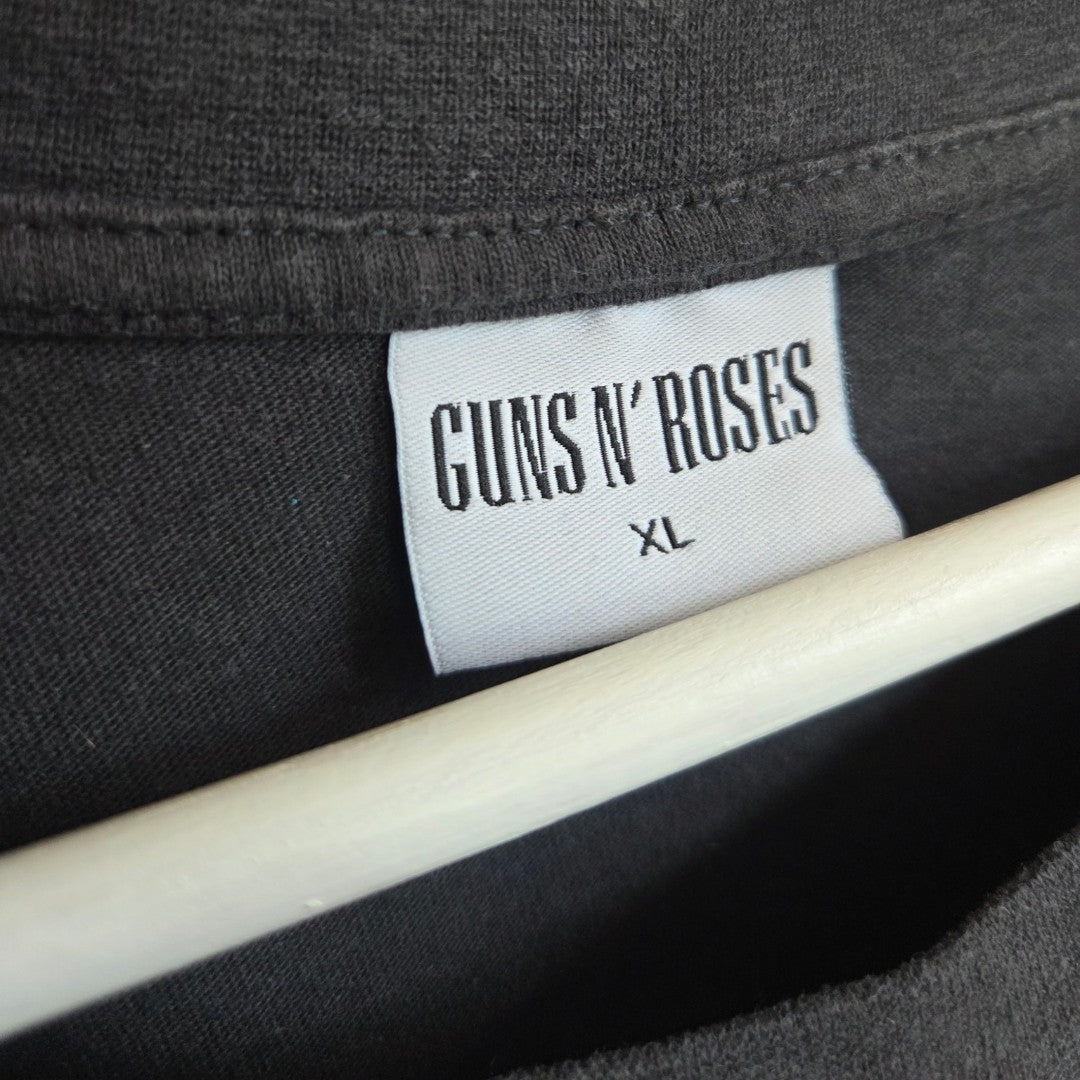 A close-up of a white label with black text inside a grey Guns N Roses Cross Band t-shirt (size XL) from Primark—upcycled for breastfeeding and pumping—emphasizes an eco-friendly, affordable option for those seeking unique and sustainable fashion. A wooden hanger is partially visible underneath.
