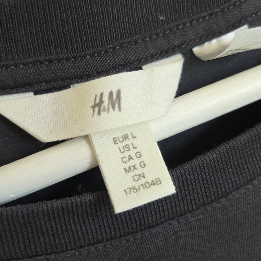 A close-up of an H&M clothing tag displaying size details—EUR L, US L, CA G, MX G, CN 175/104B—attached to the dark grey Upcycled Breastfeeding (Nursing) & Pumping Mickey Mouse t-shirt (size L, item 100533) on a white hanger.