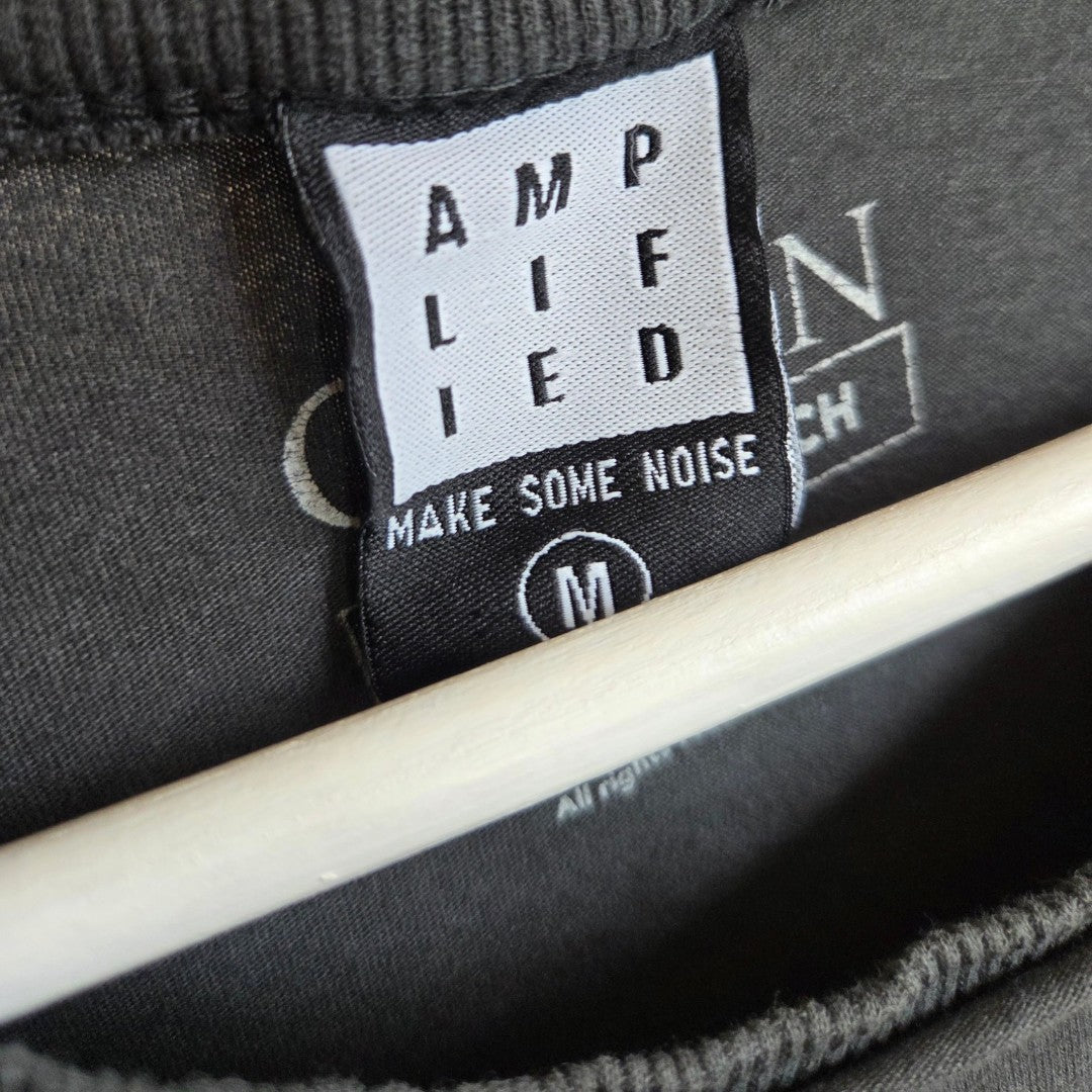 A close-up of a clothing tag on a dark shirt shows the text AMPLIFIED MAKE SOME NOISE M in black letters, with a white embroidered logo partially visible in the background, highlighting the eco-friendly Upcycled Breastfeeding (Nursing) & Pumping Queen Eagle Band t-shirt M by Amplified—perfect for those seeking sustainable style.
