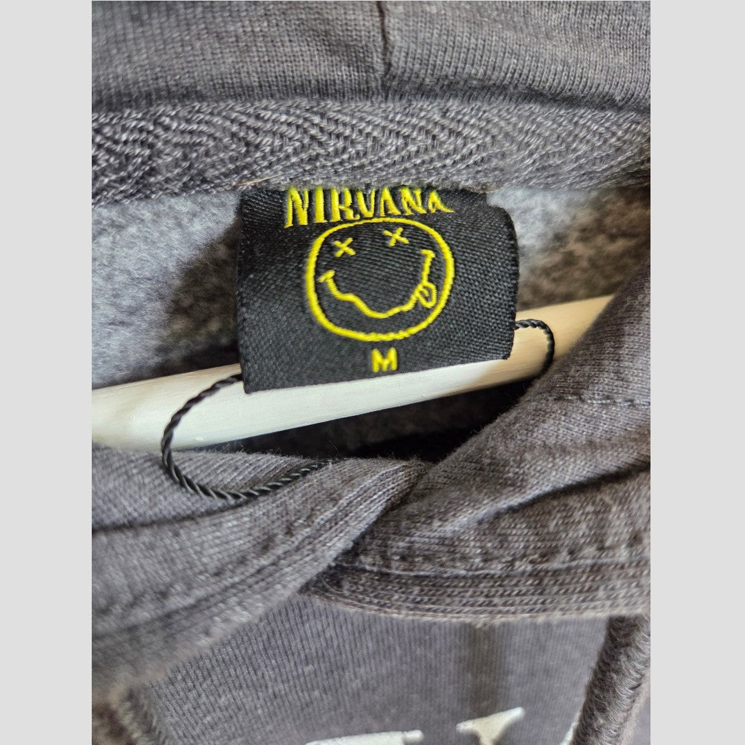 A close-up of a black clothing tag featuring the yellow Nirvana logo and smiley face, marked size M for medium, sewn inside the Upcycled Breastfeeding (Nursing) & Pumping Grey/White Nirvana Angel Band Hoodie by Primark (100521), draped over a white hanger—a stylish detail often found in this Primark nursing wear collection.