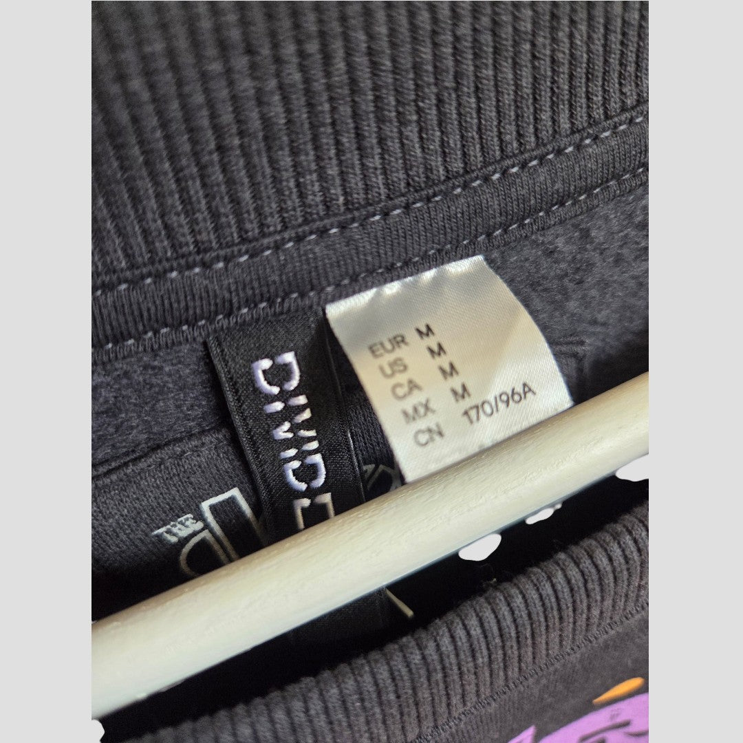 A close-up of a clothing tag on an H&M Upcycled Breastfeeding (Nursing) & Pumping The Doors Purple Band Sweatshirt M (100452) shows size M for EUR, US, CA, and MX, with a white hanger visible underneath the collar.