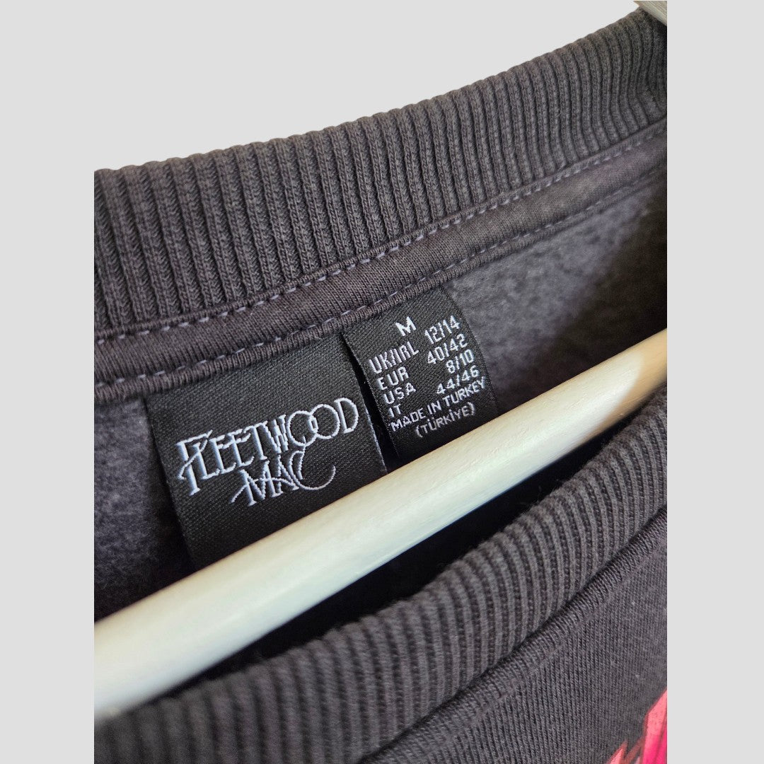 Close-up of the tag on a black Primark Upcycled Breastfeeding (Nursing) & Pumping Floral Fleetwood Mac Band Sweatshirt (size M, UK12/14, 100439), hanging on a light-colored hanger, showing size and manufacturing details.
