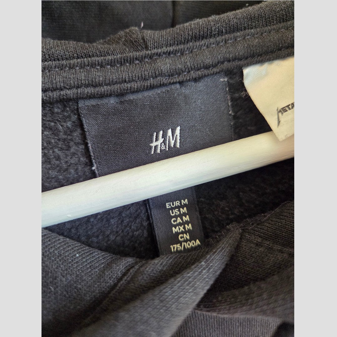 Close-up of a black H&M clothing tag hanging on a rod, displaying regional size information for the Upcycled Breastfeeding (Nursing) & Pumping Black Metallica Vertigo Band Hoodie M (100606), with a white care label partially visible—ideal for those seeking eco-friendly or nursing wear.