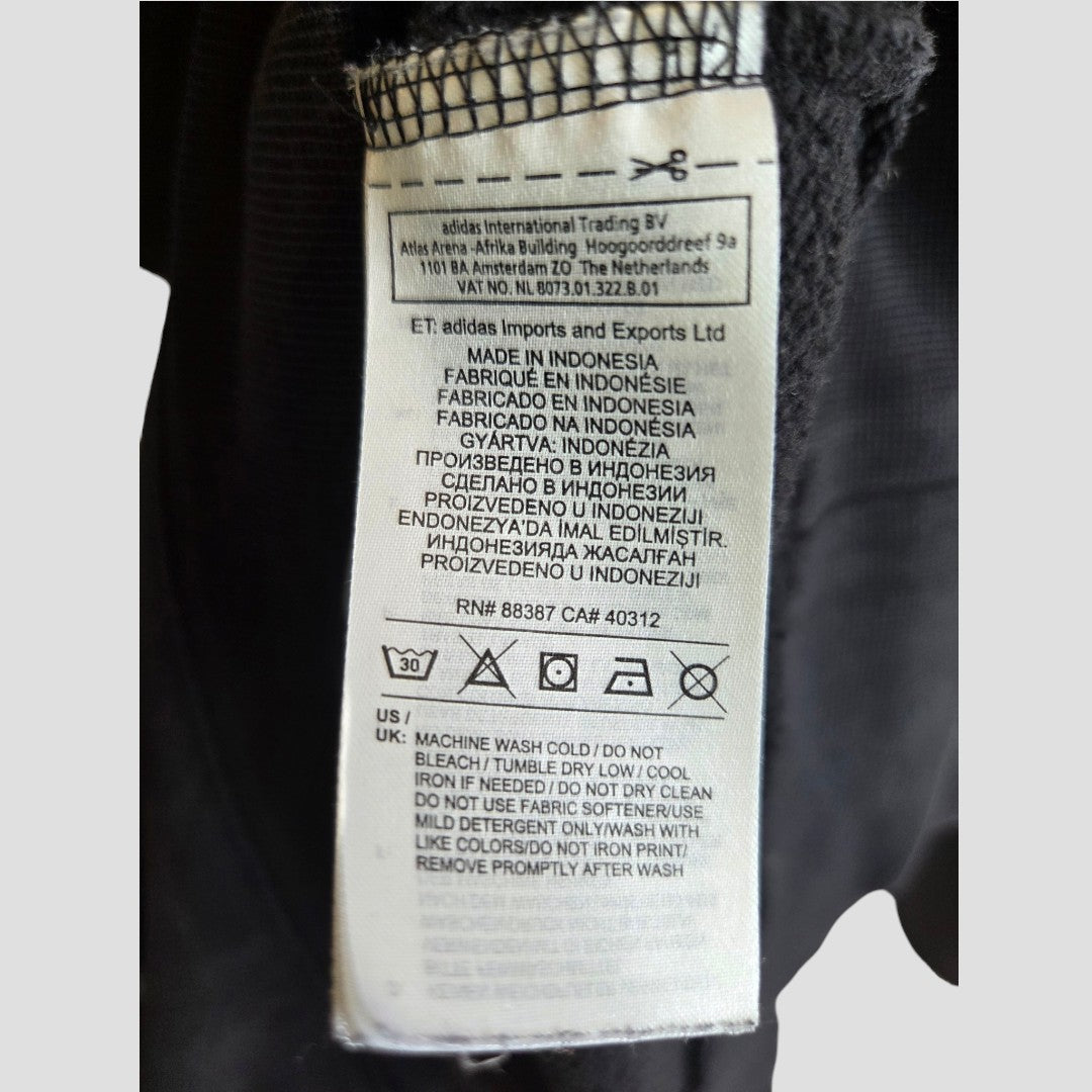 A close-up of a clothing tag on the Upcycled Breastfeeding (Nursing) & Pumping Black Adidas White Logo Hoodie UK16 (100486) by Adidas shows care instructions and manufacturing details in multiple languages, including English, Russian, and Indonesian. The tag features laundry symbols and mentions adidas Imports and Exports Ltd., underscoring the brand’s dedication to quality—making this hoodie an ideal choice for those seeking affordable, comfortable nursing wear.