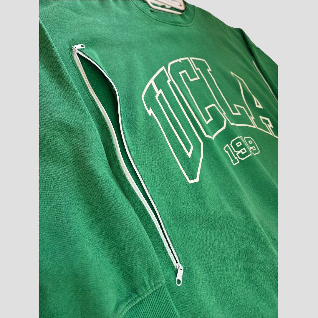 H&M’s Upcycled Breastfeeding (Nursing) & Pumping Green UCLA sweatshirt (size L, product code 100629) features eco-friendly materials and a distinctive white zippered pocket diagonally across the left chest. Large white “UCLA 199” text is printed on the front, and the angled product image highlights its textured fabric.