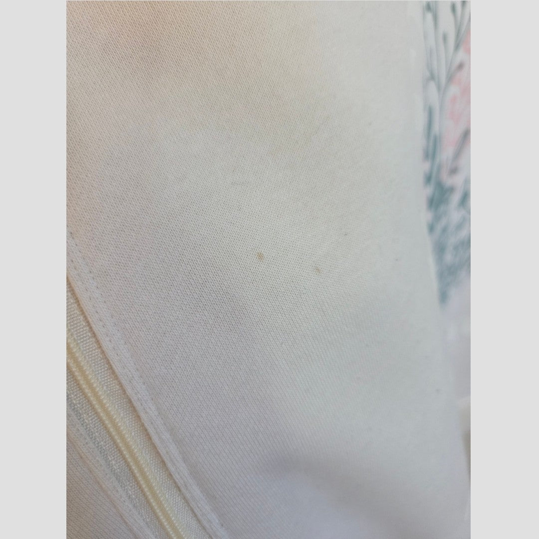 Close-up of a light-colored fabric from the Primark Upcycled Breastfeeding (Nursing) & Pumping Cream Wildflowers sweatshirt XL (100645), showing a faint yellowish area and two small brown spots. A partial floral or leafy patterned design is visible in the background on the right side, suggesting it is part of this nursing clothing item.