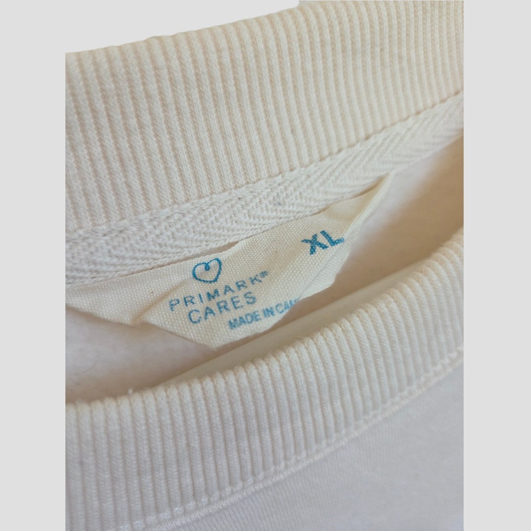 A close-up of a white clothing tag on a cream-colored Upcycled Breastfeeding (Nursing) & Pumping Cream Wildflowers sweatshirt XL by Primark shows the text “PRIMARK CARES,” “MADE IN CHINA,” and “XL” in blue. The collar and ribbed fabric are also visible—ideal for those looking for comfortable nursing or breastfeeding clothing in the Primark XL range.