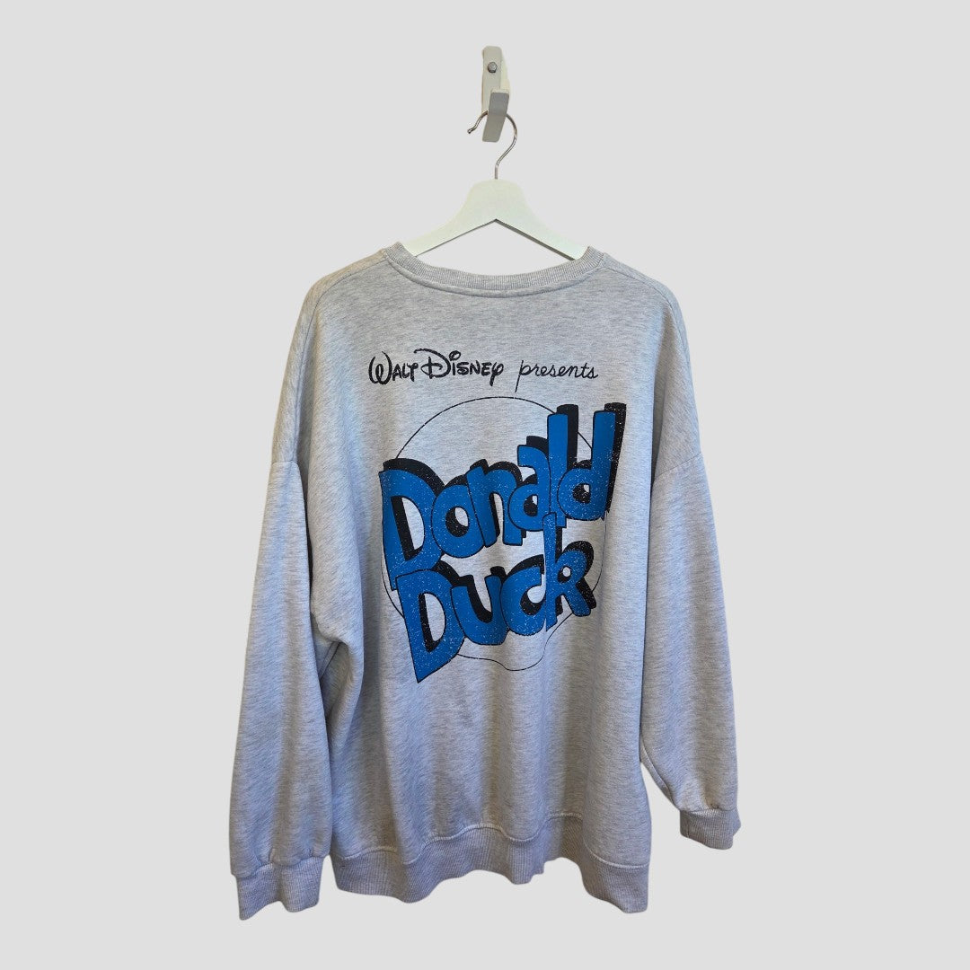 An upcycled grey Disney Donald Duck sweatshirt by Primark (size L, UK14/16) hangs on a white hanger, featuring “Walt Disney presents Donald Duck” in bold blue and black cartoon-style letters on the back—a playful and affordable addition to your nursing and pumping wardrobe.