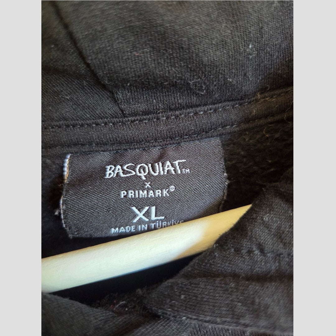 Close-up of a black clothing tag reading BASQUIAT x PRIMARK XL MADE IN TURKEY in white stitching, attached to the inside collar of a black Upcycled Breastfeeding (Nursing) & Pumping Black Basquiat Graphic hoodie XL (UK18/20) by Primark on a hanger—an eco-friendly, affordable option for stylish nursing wear.