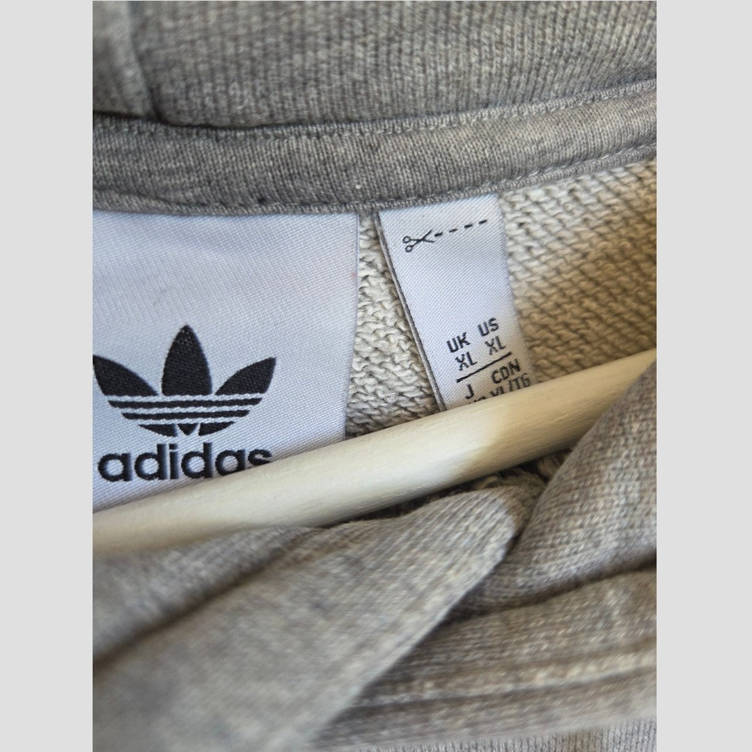 Close-up of a grey Adidas tag displaying the logo and size details in UK, US, and JPN formats on the Upcycled Breastfeeding (Nursing) & Pumping Grey/White Adidas Hoodie XL (100494). A wooden hanger is partially visible beneath the collar, emphasizing this comfortable and affordable nursing option by Adidas.