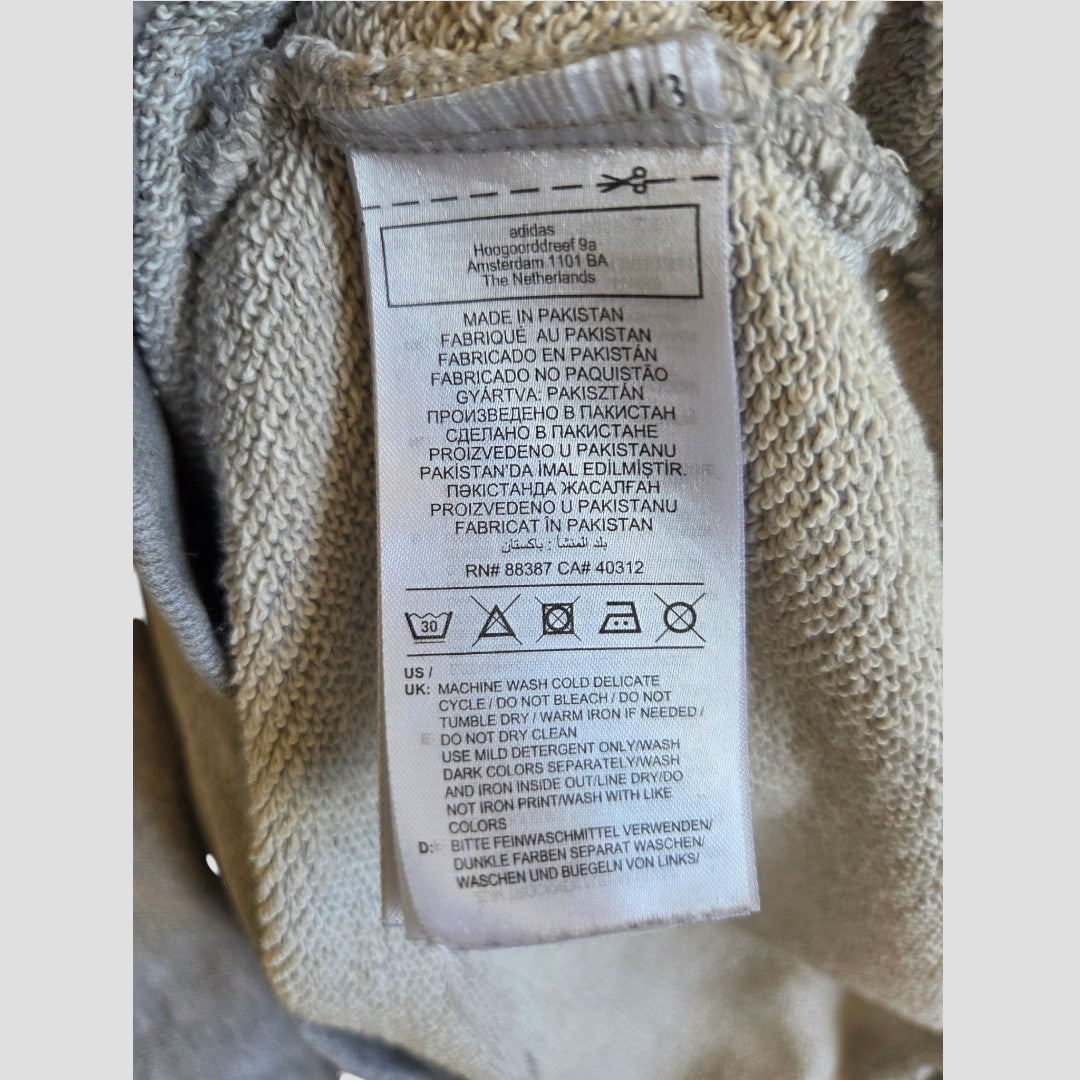 A clothing tag attached to the Upcycled Breastfeeding (Nursing) & Pumping Grey/White Adidas Hoodie XL (100494) by Adidas displays care instructions and material information in multiple languages—including English, French, and German. Designed for nursing apparel, the tag features clear washing, drying, and ironing symbols at the bottom for easy garment care.