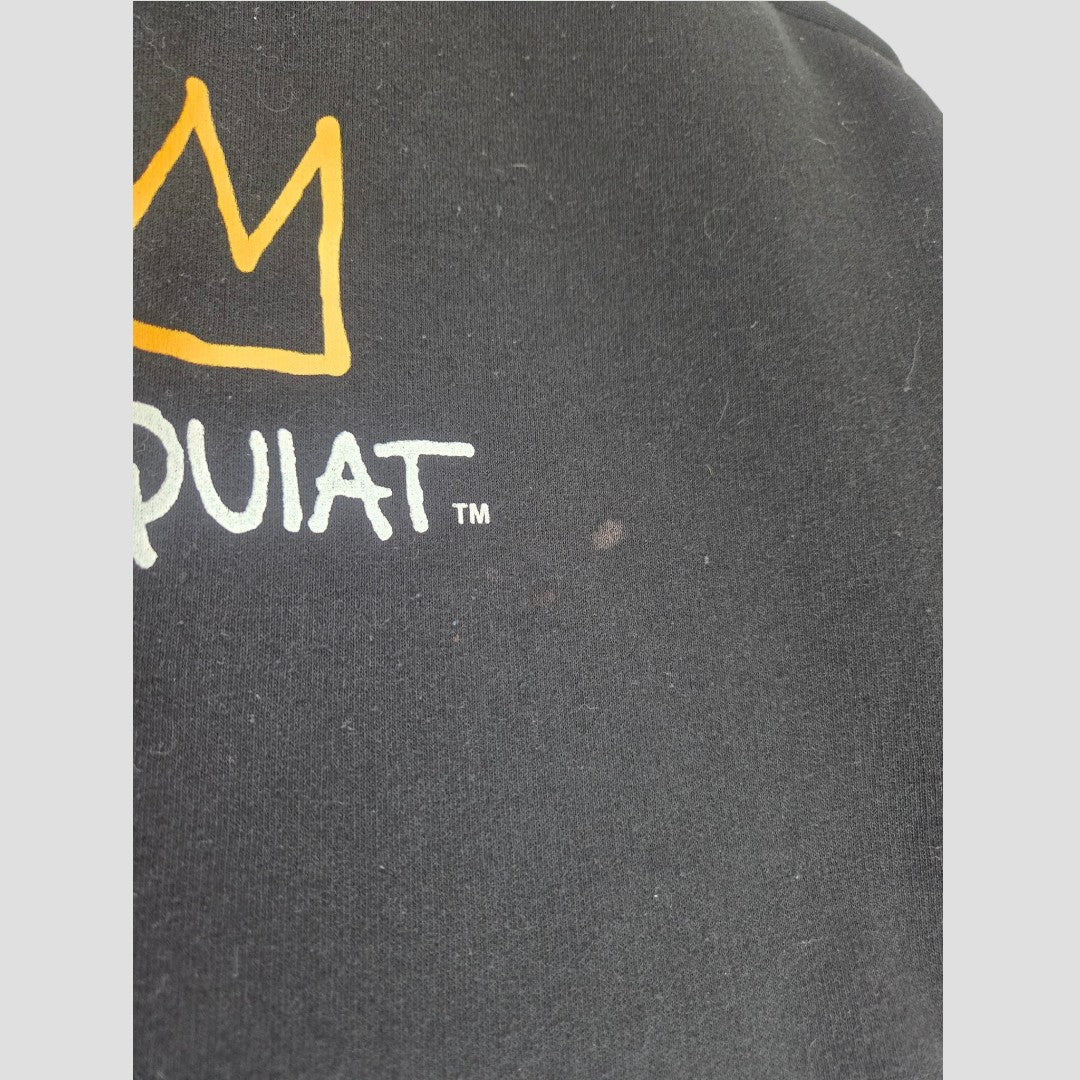Close-up of a black Primark fabric—an Upcycled Breastfeeding (Nursing) & Pumping Black Basquiat Graphic hoodie in XL (UK18/20)—showing part of the white BASQUIAT text and a yellow crown graphic. The eco affordable garment has small visible lint and a faint, round spot near the center.