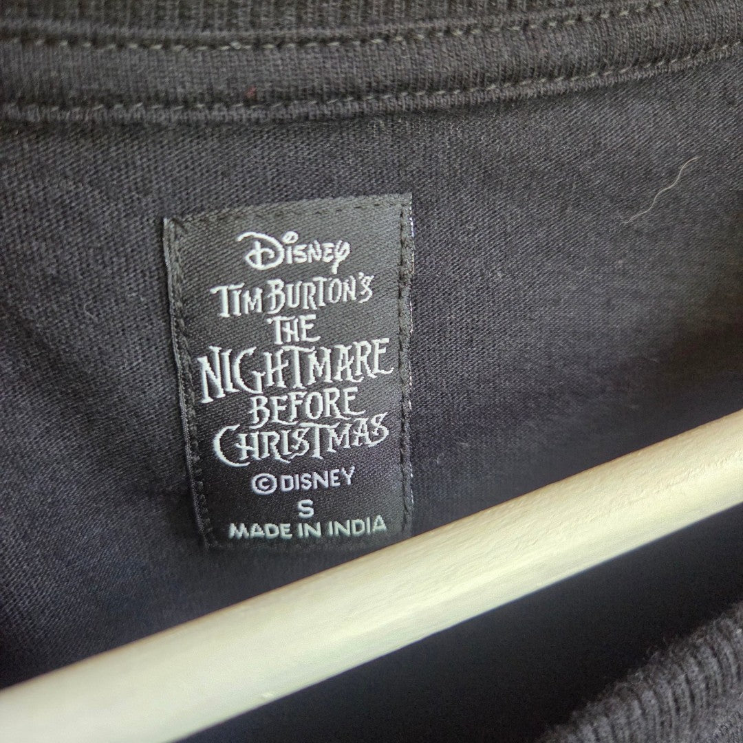 A black clothing tag on a shirt reads: Disney Tim Burton’s The Nightmare Before Christmas, S, Made in India. The shirt—an upcycled Primark Oversized Nursing & Pumping T-shirt (UK10/12)—is black and displayed on a wooden hanger. This affordable piece can be effortlessly styled with other nursing clothing essentials for versatile comfort.