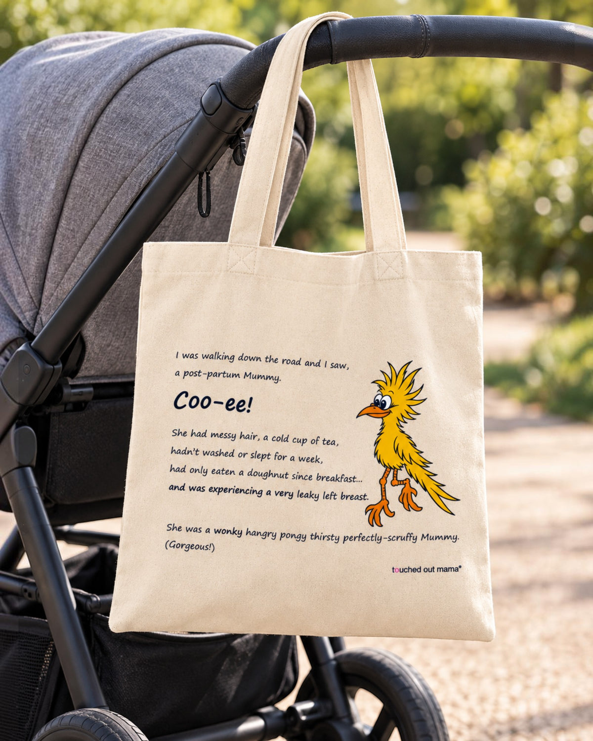 A Wonky Mummy eco tote bag hanging from a stroller handle, featuring a cartoon illustration of a yellow bird and text describing a wonky mummy.